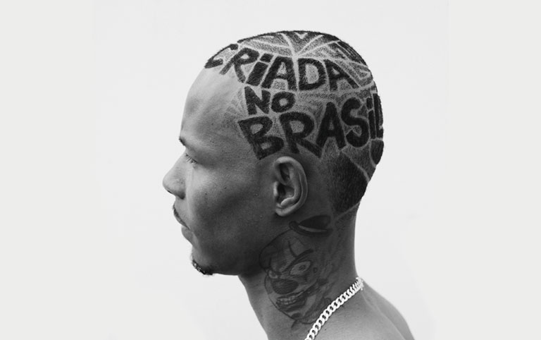 Fotografia em preto e branco de um jovem negro com o cabelo cortado com letras que formam a frase Criada no Brasil - por @caodenado - Batekoo Festival